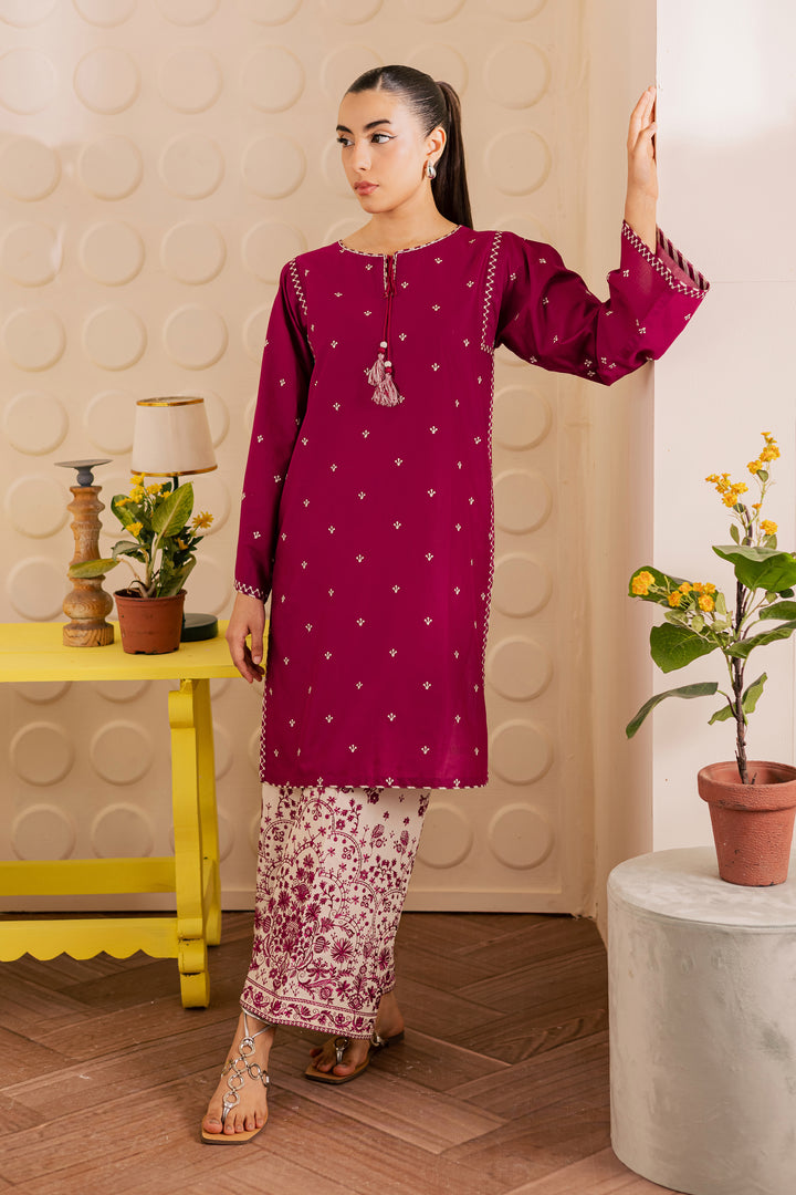Woman wearing a magenta embroidered top and white pants with pink patterns in a room with decorative elements.