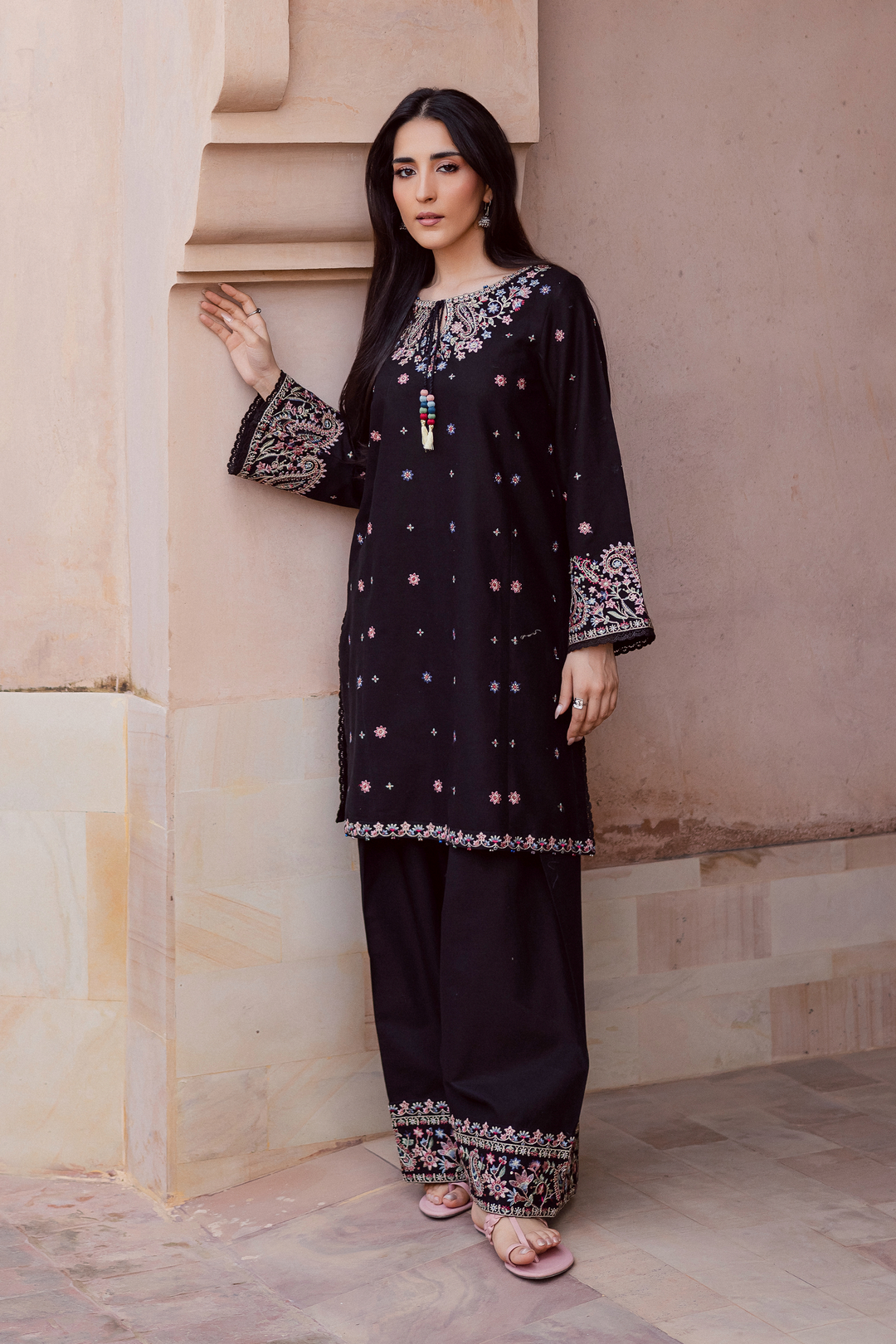 Black embroidered two-piece kurta set for women featuring colorful floral embroidery on sleeves and hems, paired with wide trousers and sandals. Traditional Pakistani outfit ideal for festive or casual wear