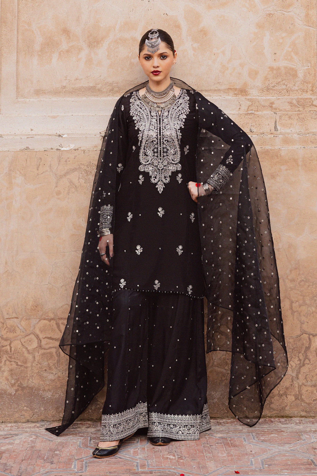 Batik UAE Arbish three piece black raw silk set featuring an embroidered paneled short shirt with crystal detailing paired with an embroidered raw silk sharara and organza dupatta. Affordable ethnic South Asian Desi Indian Pakistani designer wear UAE fashion for teens adults girls and women available in sizes XS S M L XL.